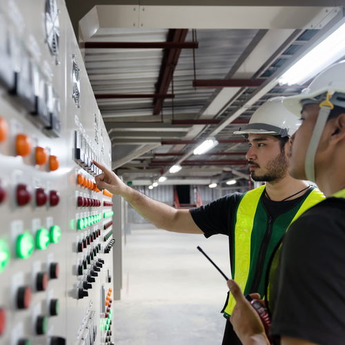 Workers wearing white helmets and yellow high-visibility vests stand in front of a control cabinet