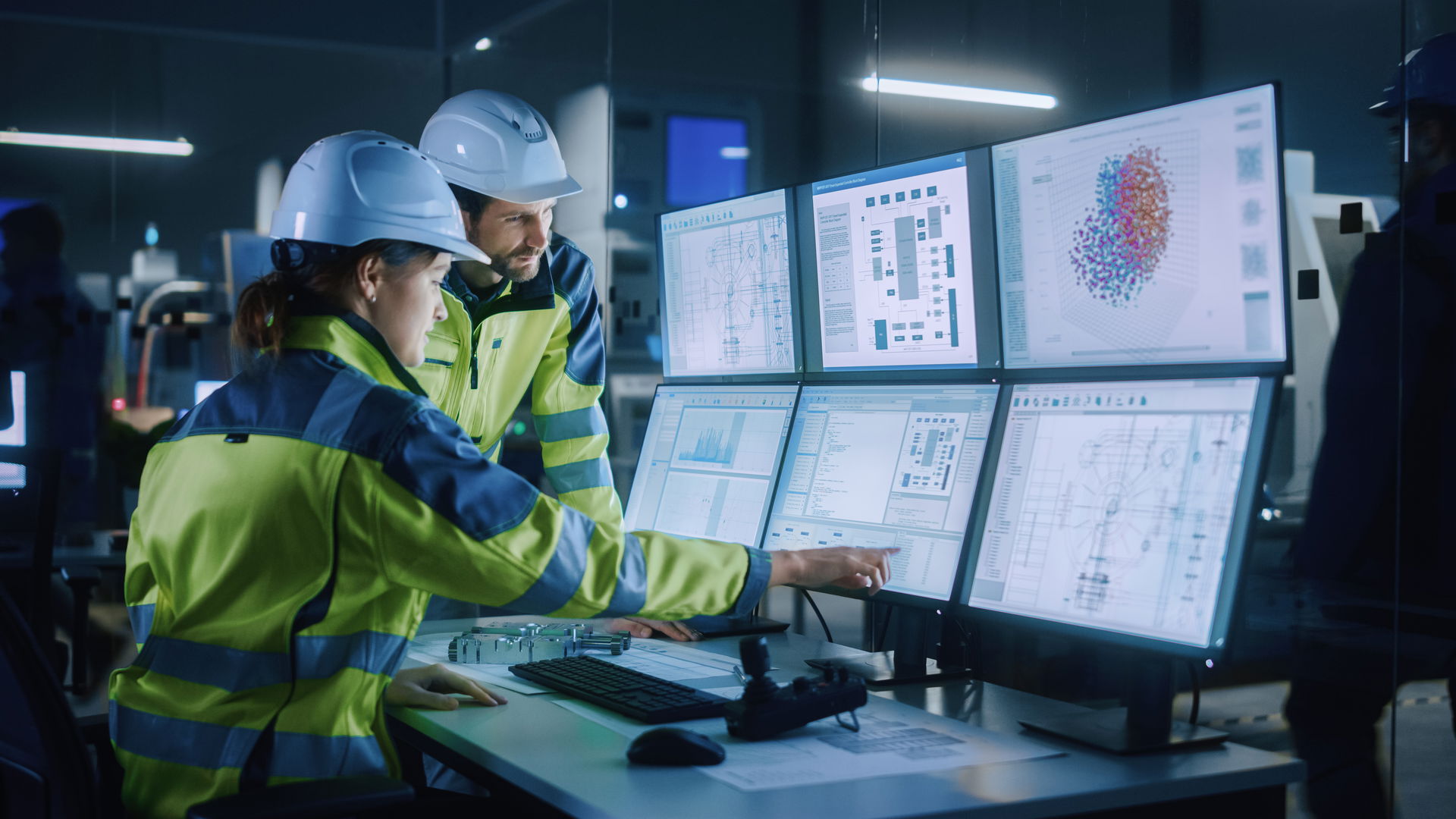 Workers in yellow work clothes and white helmets in front of a monitor wall
