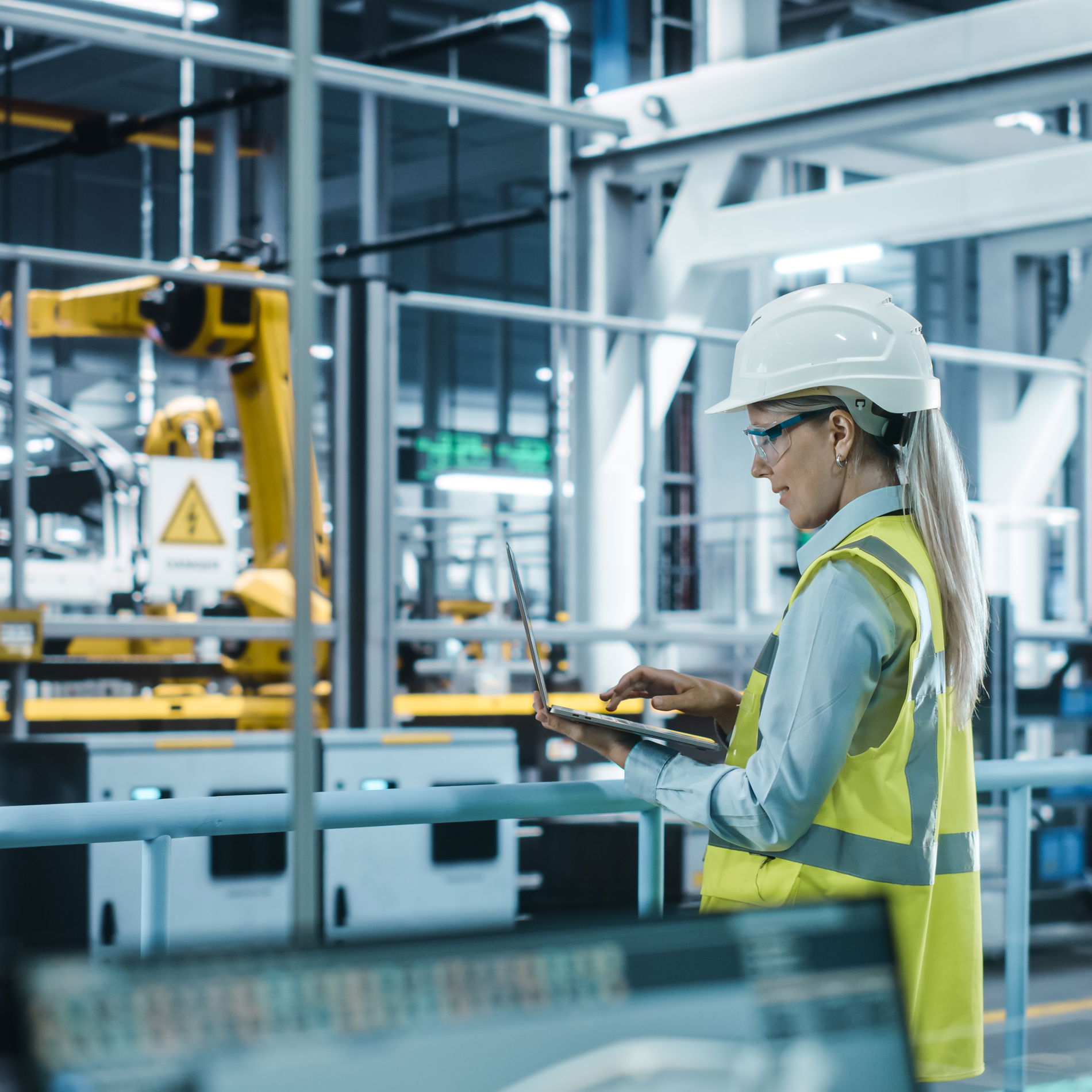 Worker wearing a helmet and vest in the office in front of a glass wall facing the production area and robot arm