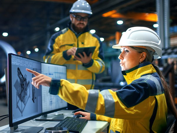 Woman and man in yellow work clothes and helmets in front of a screen
