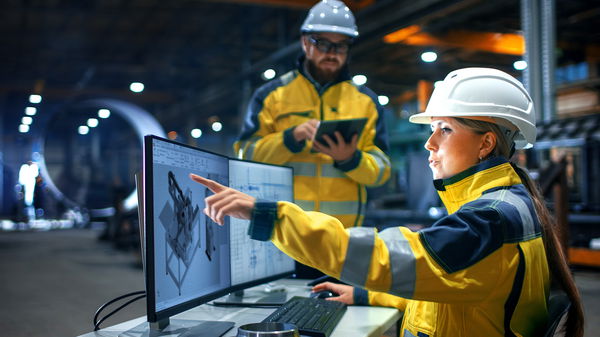 Woman and man in yellow work clothes and helmets in front of a screen
